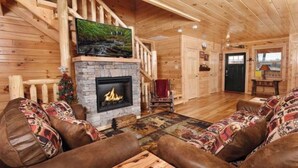 Ein Holzblockhaus-Interieur mit Kamin aus Stein, Holzwänden, einem darüber angebrachten Fernseher und einer gemütlichen Sitzecke mit gemusterten Kissen.