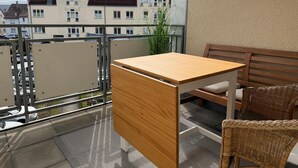 En balkon med et træbord og fletstole, med udsigt over et boligområde med huse og en klar himmel.