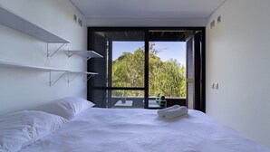 Quarto de hotel moderno com uma cama grande, varanda com vista para árvores e uma pequena mesa com uma cadeira.