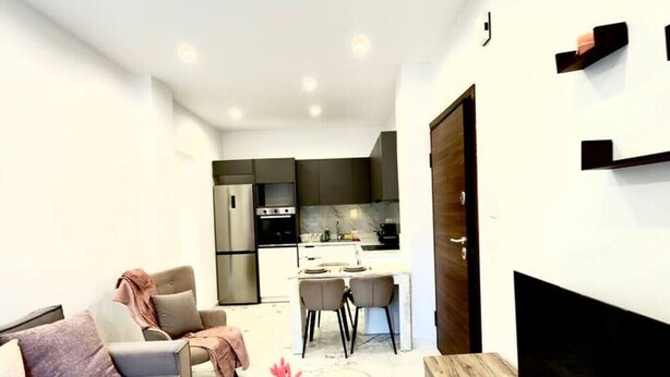 Una sala de estar moderna con sofá, mesa de centro y televisor. Se observa una cocina al fondo con zona de comedor.