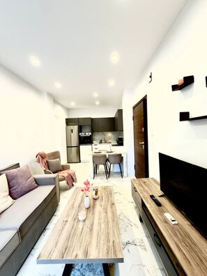 Un salon moderne avec un canapé, une table basse et un téléviseur. Au fond, on aperçoit une cuisine avec un coin repas.