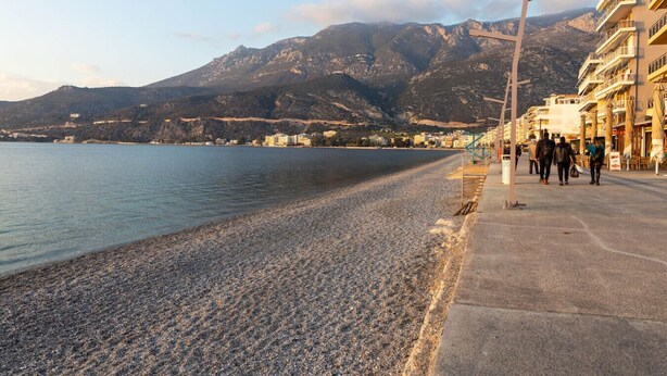 A pebble beach with a mountain backdrop, a pedestrian path, and buildings along the shore.