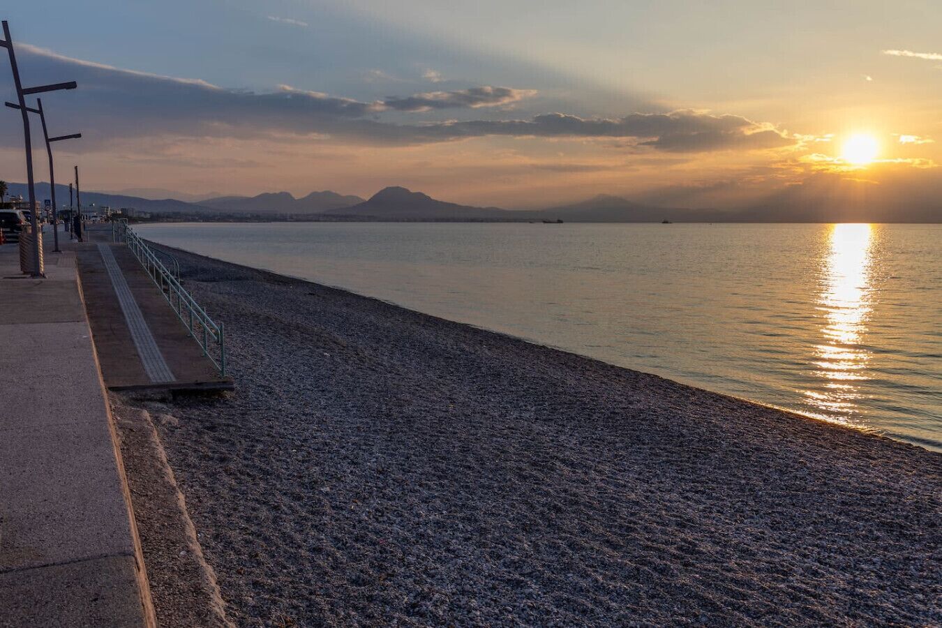 A waterfront promenade with a paved walkway, metal railings, and distant mountains.