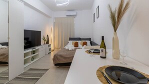 A modern hotel room with a bed, a TV, a minibar, a dining area with a bottle of wine and glasses, and a large window.