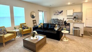 Una sala de estar moderna con un sofá negro, sillones amarillos y una mesa de centro. Hay una zona de cocina con un refrigerador y un lavamanos.