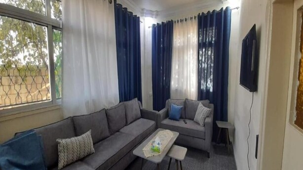 A living room with a grey sofa, a small table, and a TV mounted on the wall. - Homey Apt in Jerash w/ City-View. Explore Jerash Ruins from this Spacious Apt (Jerash)