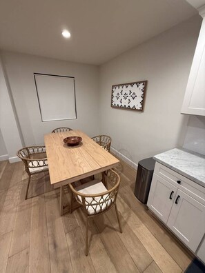 Ein Essbereich mit Holztisch, Korbstühlen, einem weißen Schrank mit Schubladen und einem Mülleimer.