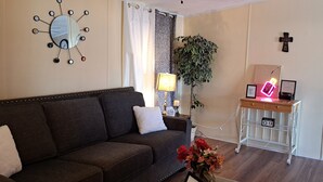 Un salon avec un canapé marron foncé, une horloge murale, une lampe, une plante, une enseigne ancienne et une petite étagère blanche sur laquelle est posé un cadre photo.