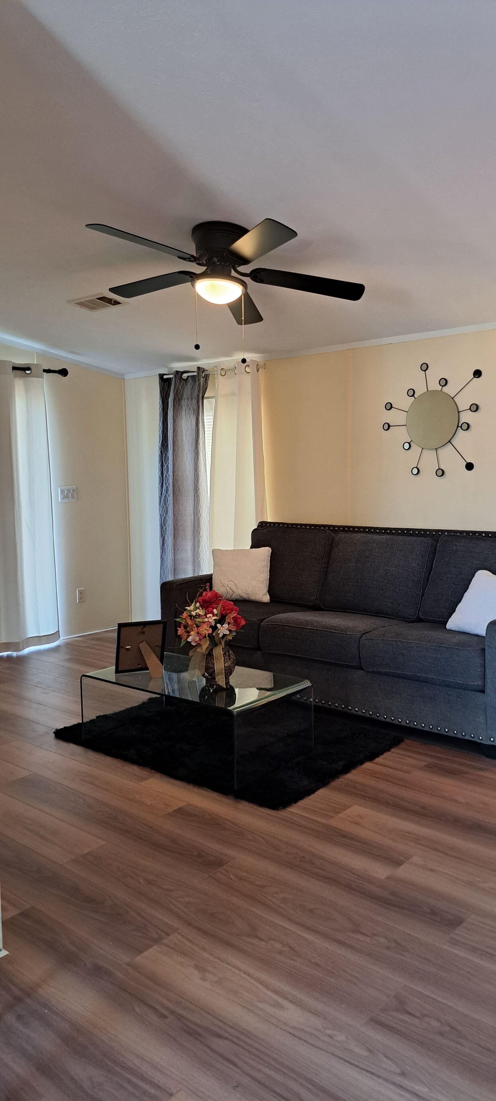 A modern living room with a ceiling fan, a dark grey sofa, a glass coffee table with a vase of flowers, and a wall-mounted decorative piece.