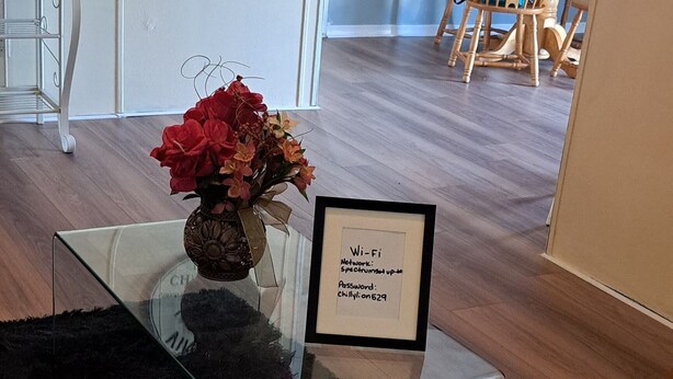 Un salon avec une table basse en verre, un vase rempli de fleurs, un panneau encadré indiquant le mot de passe du Wi-Fi et une horloge murale ancienne.