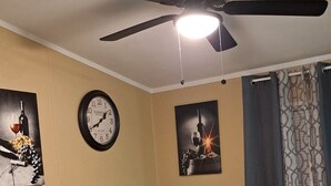 Un coin repas comprenant une table ronde en bois et quatre chaises, un ventilateur de plafond, une horloge murale et deux tableaux encadrés.