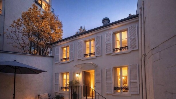 Un edificio de dos pisos con paredes y contraventanas blancas, una escalera que conduce a la entrada, y un pequeño espacio al aire libre con una mesa y sillas.