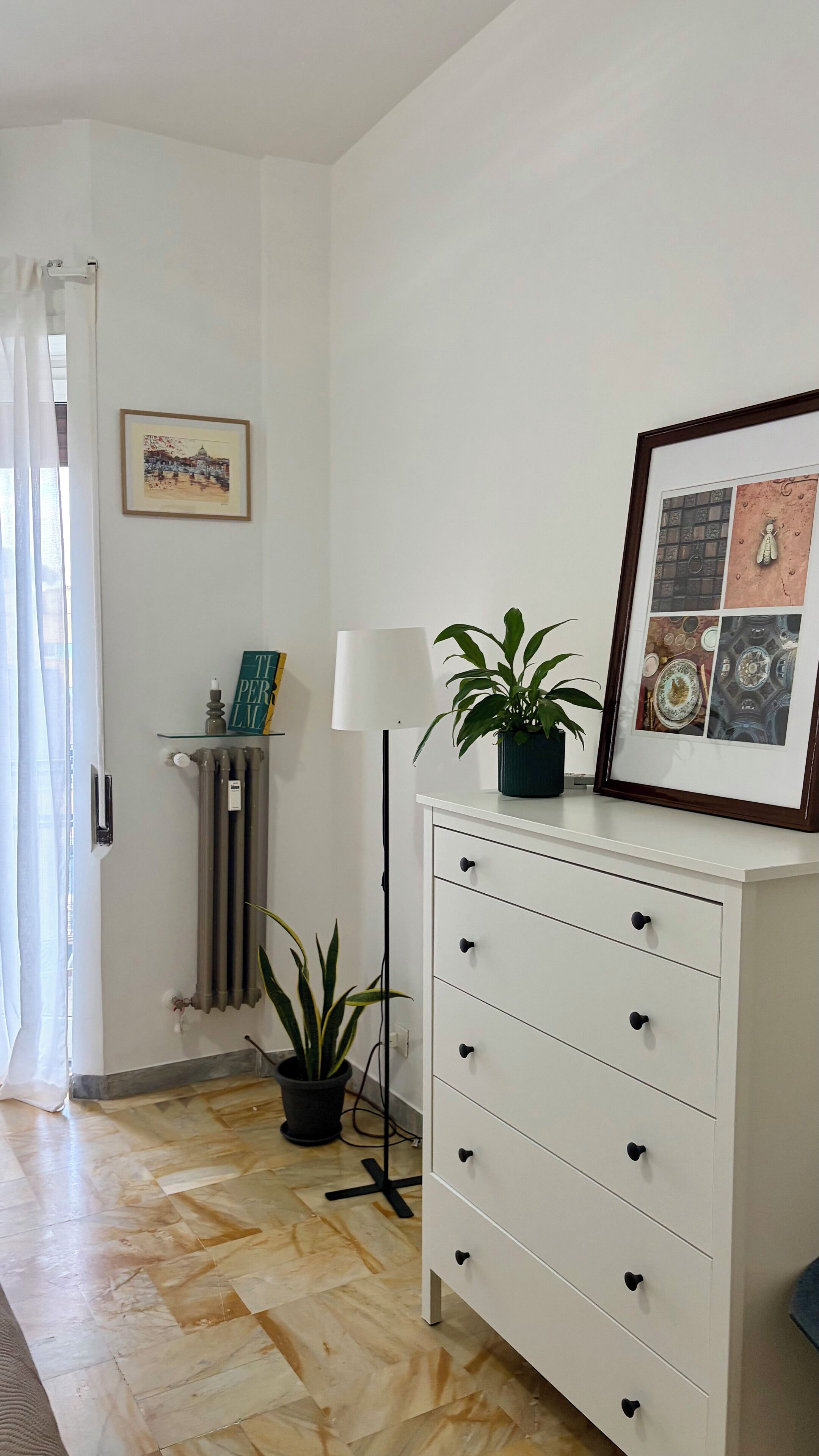 Une pièce avec une commode blanche, un tableau encadré, une plante en pot, un radiateur et une fenêtre avec des rideaux.