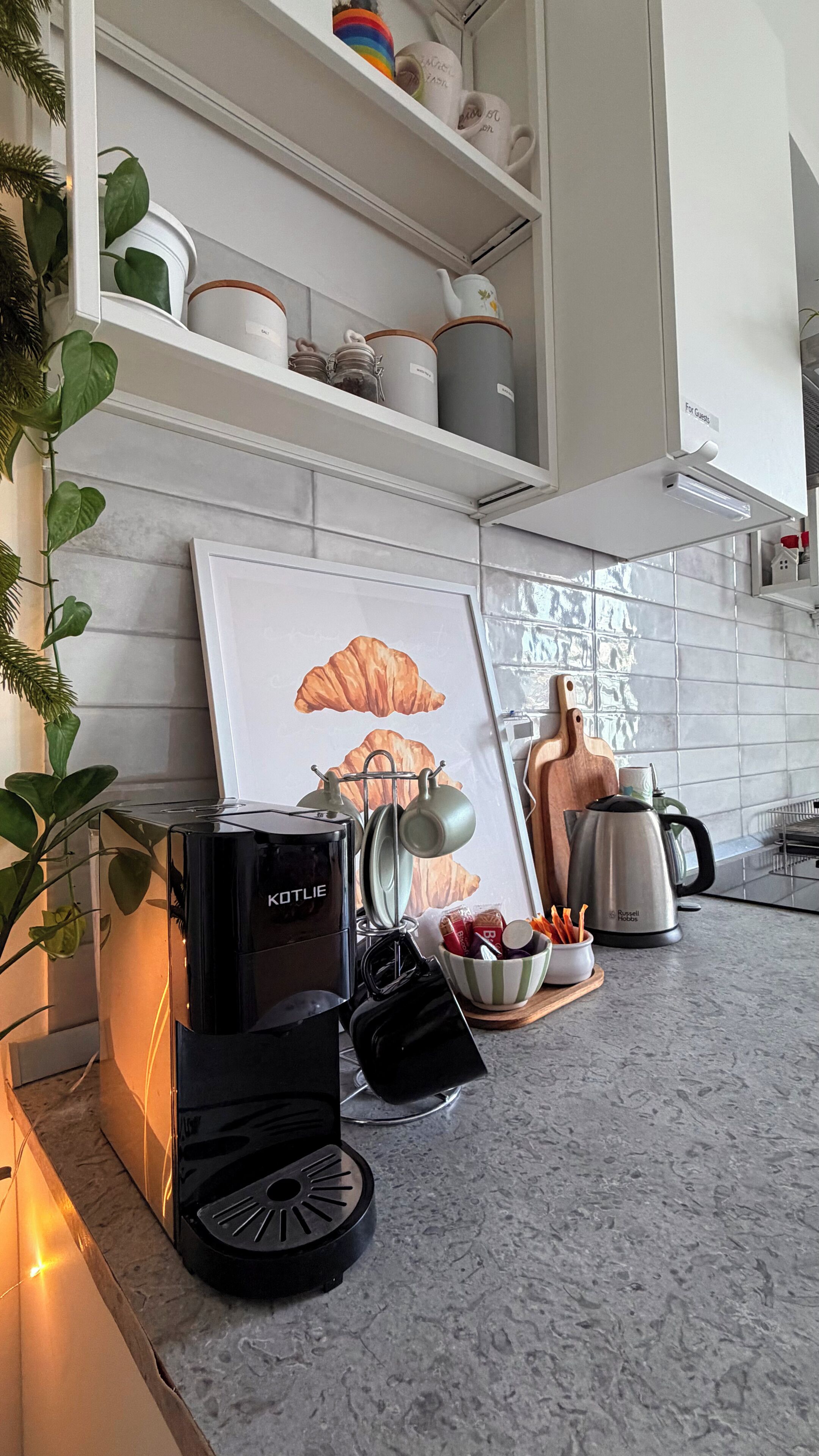 A kitchen with white cabinets, a framed picture of croissants, a kettle, a coffee maker, and a bowl of utensils.