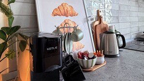 Una cocina con armarios blancos, un cuadro con cruasanes, una tetera, una cafetera y un bol de utensilios.