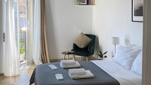 Una habitación de hotel con cama, una silla, una mesita, una mesita de noche, una lámpara, un marco de fotos y una luz en el techo.