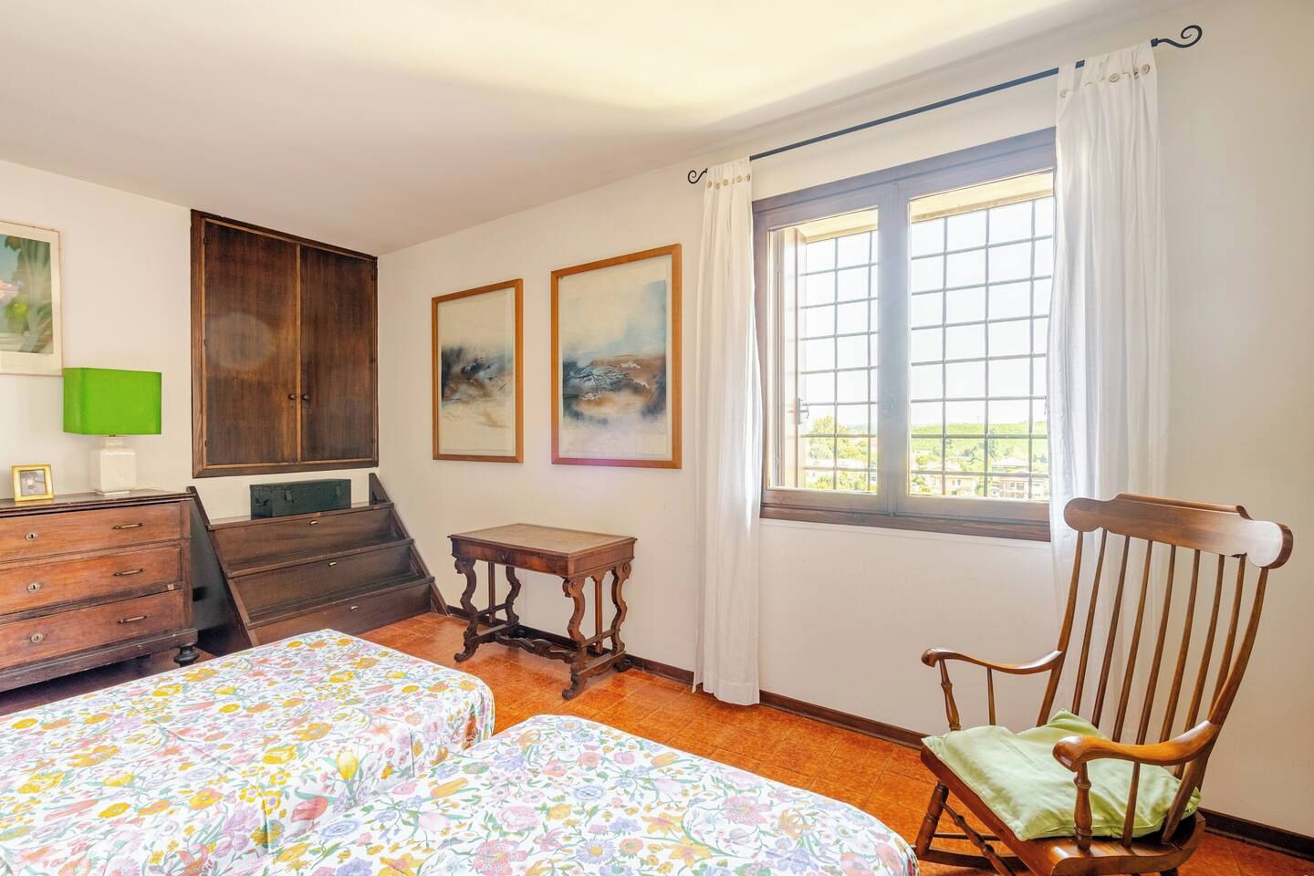 A bedroom with two beds, a wooden chair, a small table, a dresser, and a window with curtains.