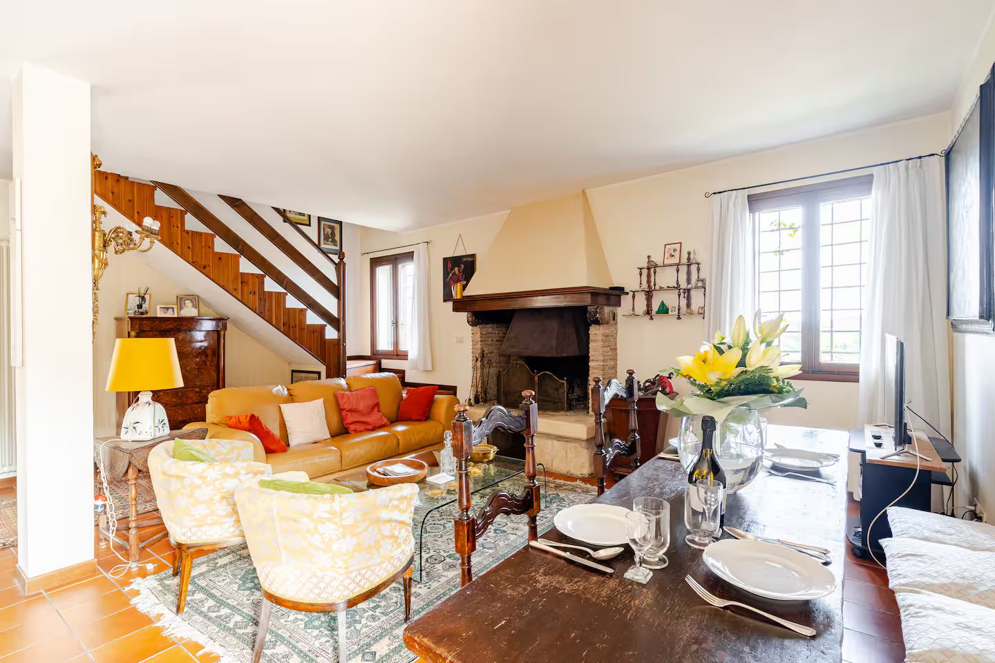 A living room with a sofa, chairs, a dining table set for two, a fireplace, and a staircase.