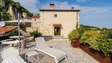A stone building with a tiled roof, a door, and a small window. There are outdoor seating areas with chairs and tables.