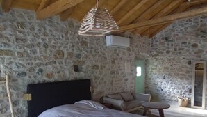 Een slaapkamer met een stenen muur, houten plafond, een bed, een stoel en een kleine tafel.