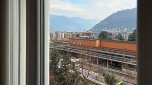 Une vue à travers une fenêtre sur un paysage urbain, avec des bâtiments, une voie ferrée et des montagnes en arrière-plan.