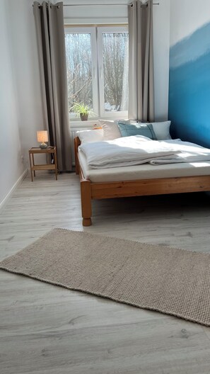 Une chambre à coucher avec un lit en bois, une petite table de chevet, une lampe, une fenêtre avec des rideaux et une vue sur les arbres à l’extérieur.