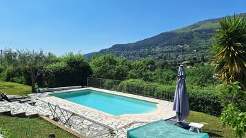 Une piscine aux bords recouverts de pierres, entourée d’une clôture métallique, et offrant une vue sur une colline verdoyante avec des bâtiments en arrière-plan.
