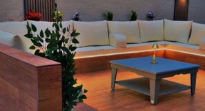 Un área de estar moderna al aire libre con un sofá y una mesa, rodeada de plantas y un banco de madera.