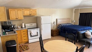 A compact kitchen with a stove, sink, and cabinets, a dining area with a table and chairs, and a bed with a blue blanket.