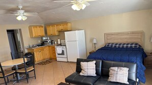 A compact room with a kitchenette, a dining area with a table and chairs, a sofa with pillows, and a bed with a blue blanket.