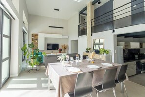 Un'elegante zona pranzo con un tavolo lungo apparecchiato per sei persone, circondato da sedie. Si intravede sullo sfondo una cucina con un piano di lavoro nero e elettrodomestici in acciaio inossidabile.