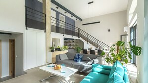 Een moderne woonkamer met een grijze bank, een witte salontafel en een trap naar een hoger gelegen verdieping.