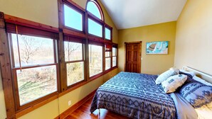 Une chambre avec une grande fenêtre, un lit recouvert d’une literie à motifs et un tableau accroché au mur.