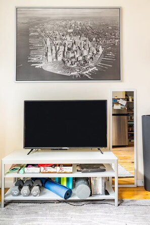 Ein Wohnzimmer mit einem Flachbildfernseher auf einem weißen Schrank, einem Bücherregal mit verschiedenen Gegenständen und einem großen schwarz-weiß Stadtbild über dem Fernseher.
