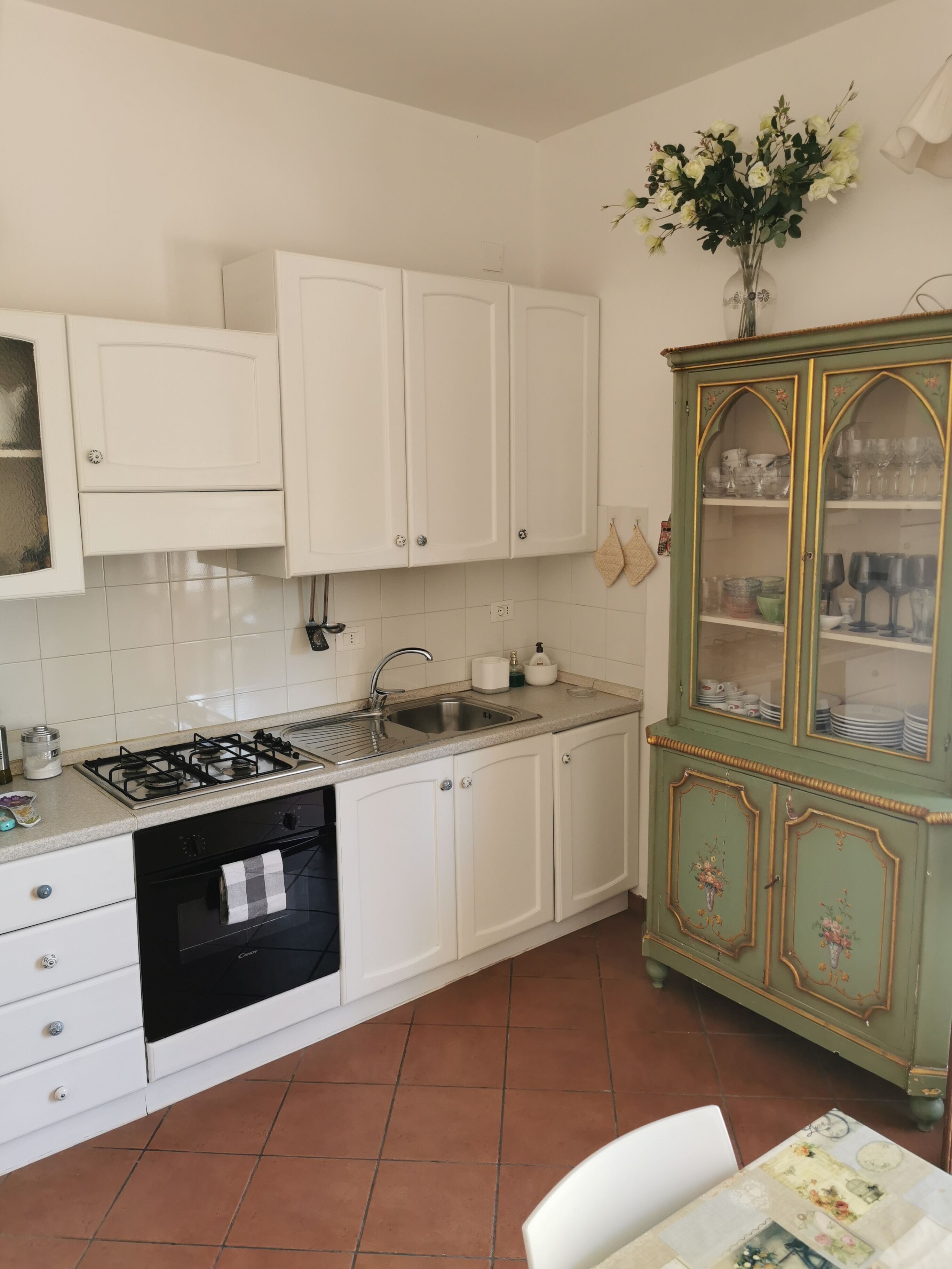Una cocina con armarios blancos, estufa a gas, lavaplatos y un fregadero. Hay un mueble verde con puertas de vidrio que contiene vajilla.