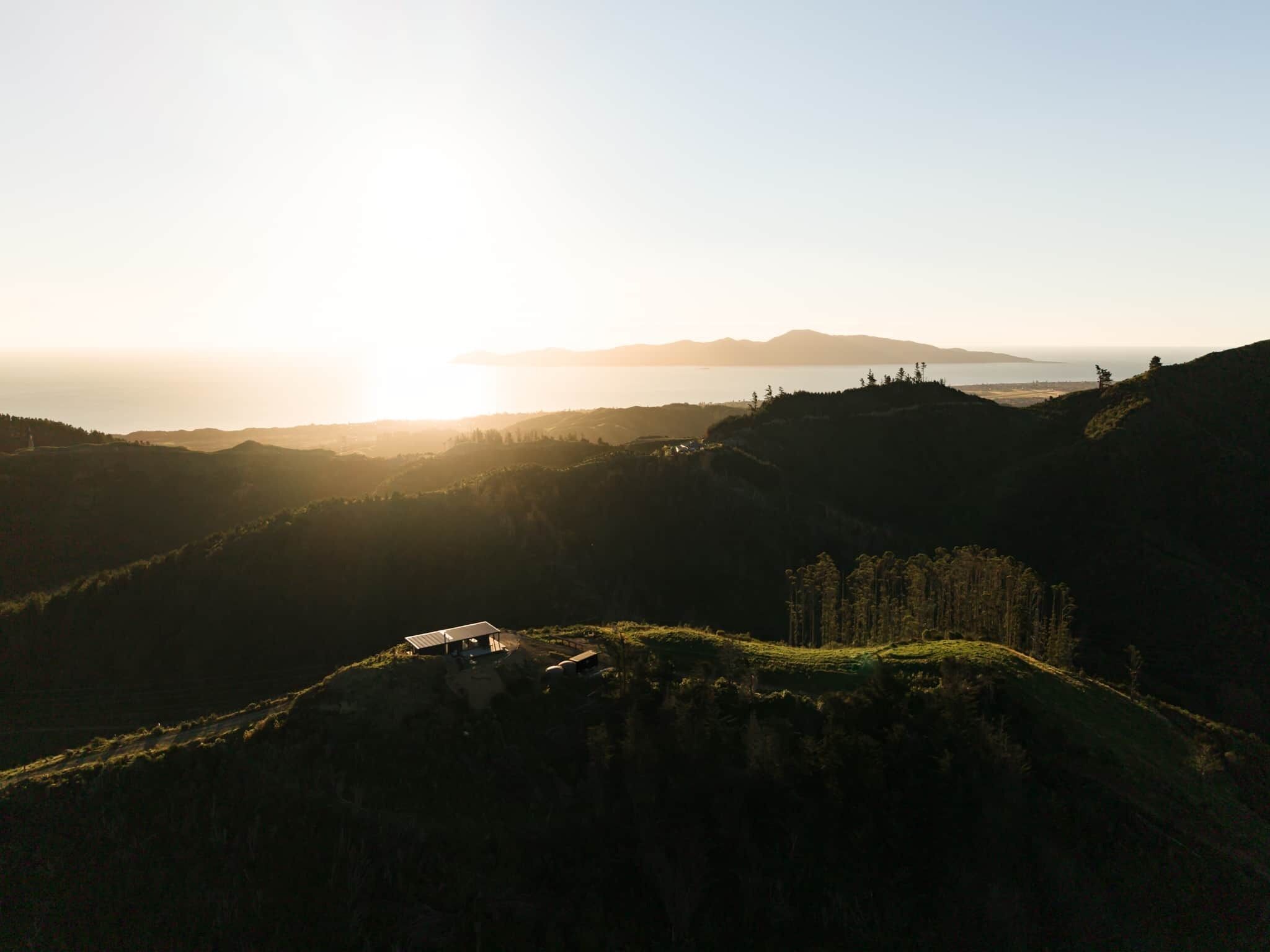 A mountainous landscape with a small building on a hill, overlooking a body of water and distant hills.