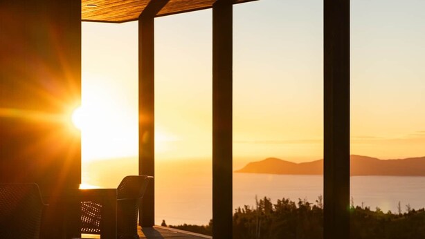 A wooden deck with chairs, a pergola, and a view of the ocean at sunset. - The Hide Kapiti Accommodation (Maungakotukutuku)