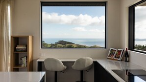 A room with a large window, two bar stools, a bookshelf, and a wine bottle. - The Hide Kapiti Accommodation (Maungakotukutuku)