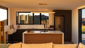 A modern kitchen with a large window, a dining area with a white table, and a sofa. - The Hide Kapiti Accommodation (Maungakotukutuku)
