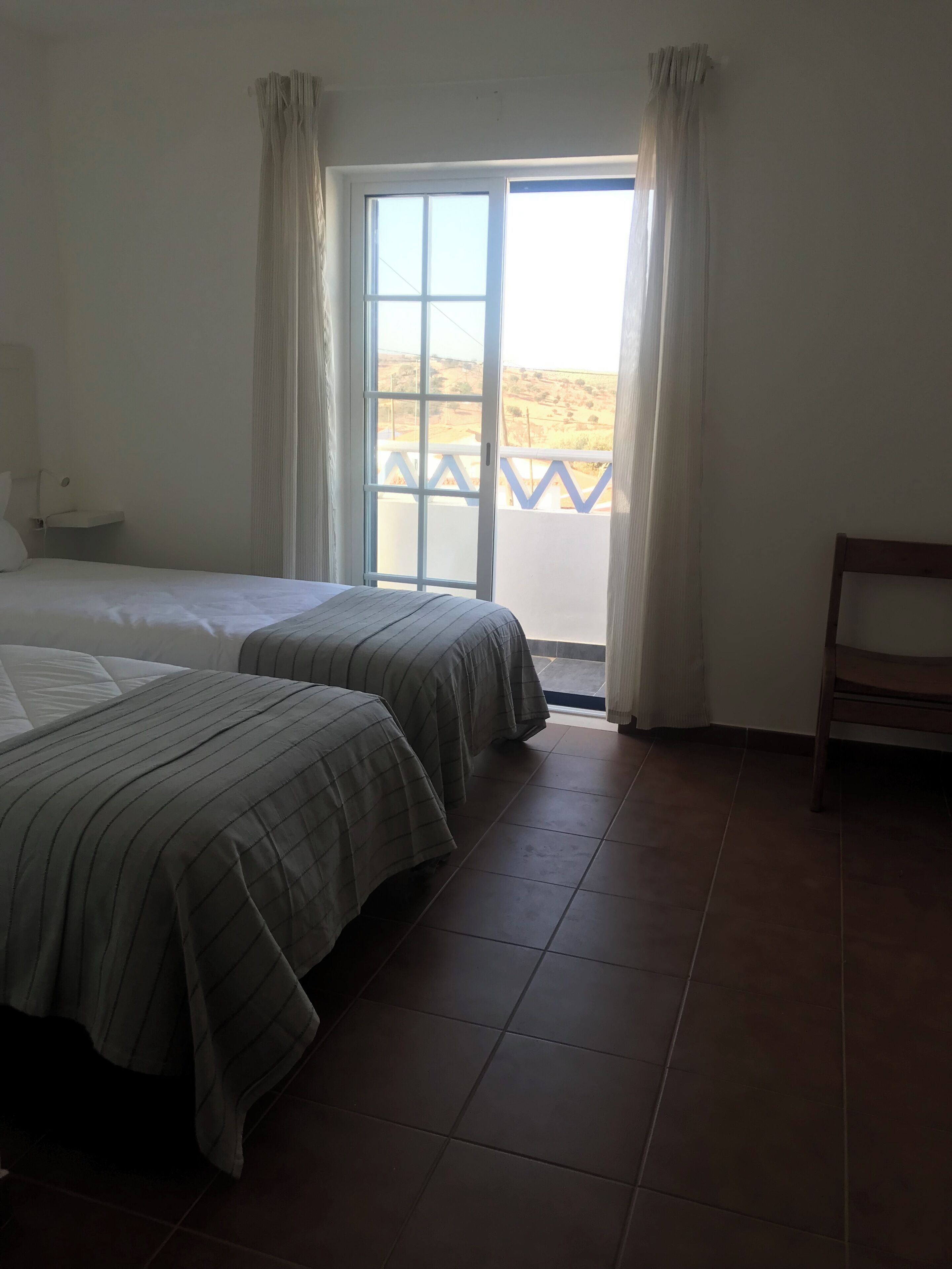 A hotel room with two beds, a bench, a window with curtains, and a view of a landscape.