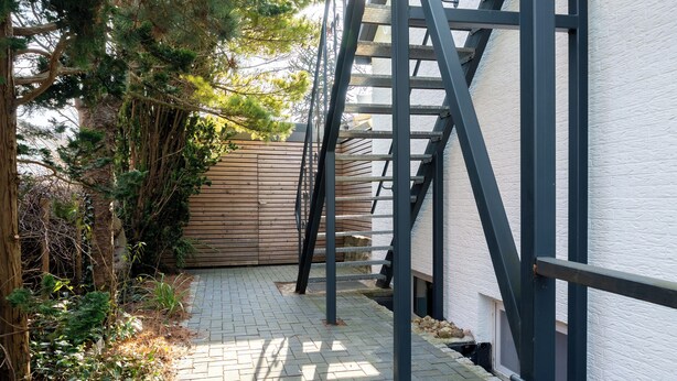 A narrow outdoor pathway leading to a building with a wooden fence and metal staircase. - Vacation apartment MeerZeit (Stein)