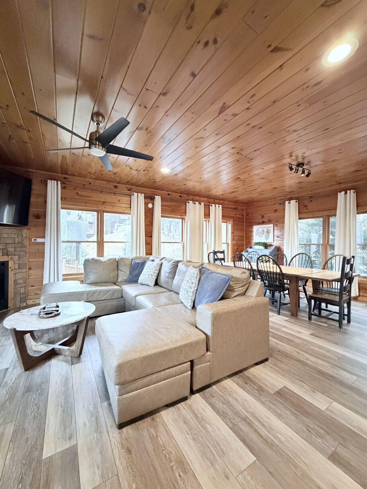 A cozy cabin interior with a wooden ceiling, a sectional sofa, a dining table with chairs, and a fireplace.