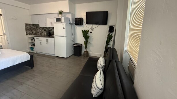 Ein kompakter Wohnbereich mit Küchenzeile, einem flachbildfernseher an der Wand, einem schwarzen Sofa mit Zierkissen und einem Fenster mit Jalousien.