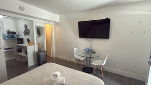 Ein Hotelzimmer mit einem Bett, einem kleinen Essbereich mit einem Glastisch und weißen Stühlen, einem an der Wand montierten Fernseher und einer Küchenzeile mit einer Mikrowelle und einer Kaffeemaschine.