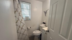 Ein kleines, sauberes Badezimmer mit Toilette, Waschbecken und Duschvorhang. Es liegen Pflegeprodukte am Waschbecken und ein Handtuch auf der Toilette.