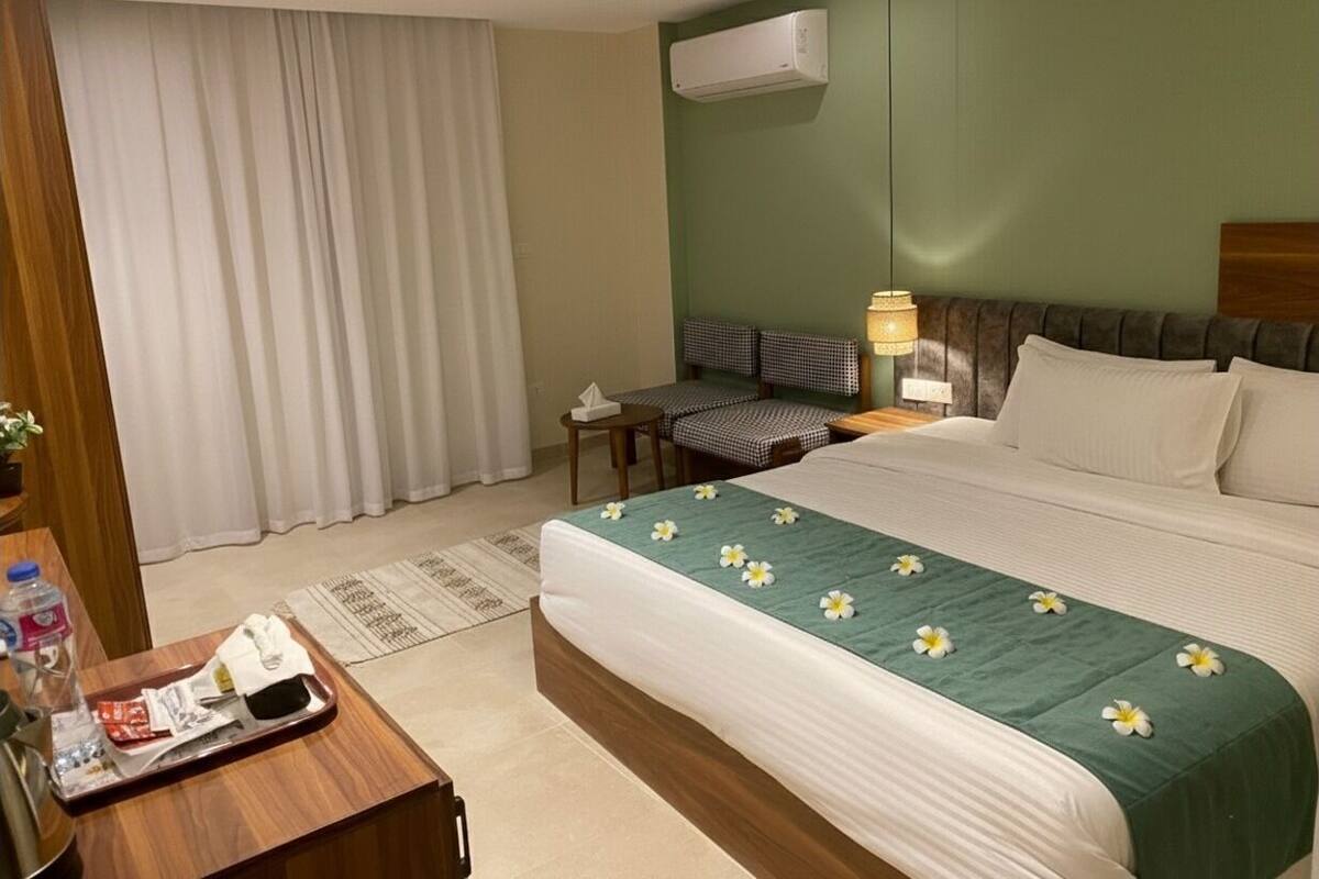 Una habitación de hotel con cama, escritorio, silla, mesita de noche, aire acondicionado y ventana con cortinas.