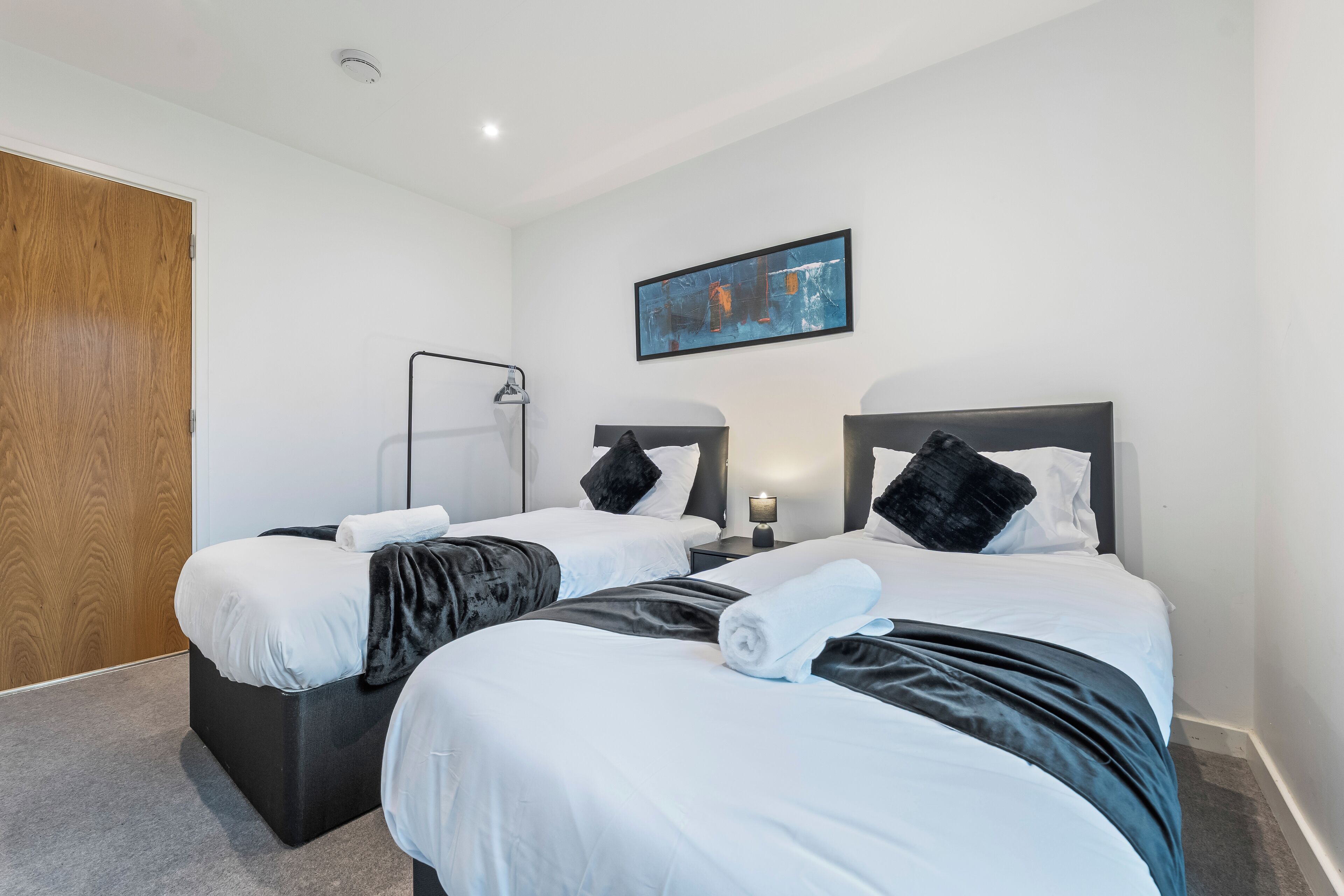 A hotel room with two beds, a wooden door, a painting on the wall, and a nightstand with a lamp.