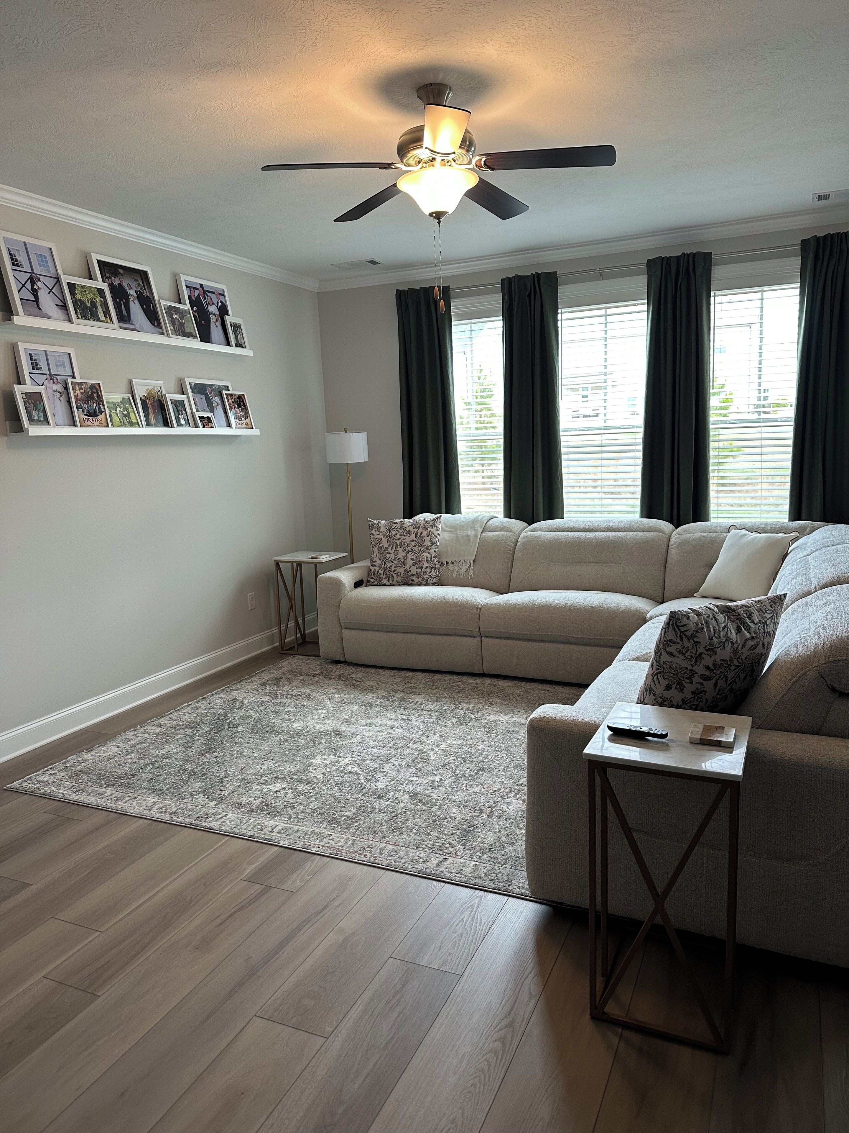 A living room with a beige sofa, a coffee table, a floor lamp, and a ceiling fan. There are framed pictures on the wall and a window with curtains.