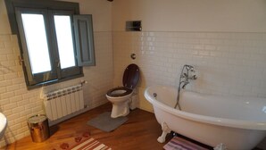 Une salle de bain avec une baignoire blanche, des toilettes en bois, une fenêtre avec des volets et un radiateur.