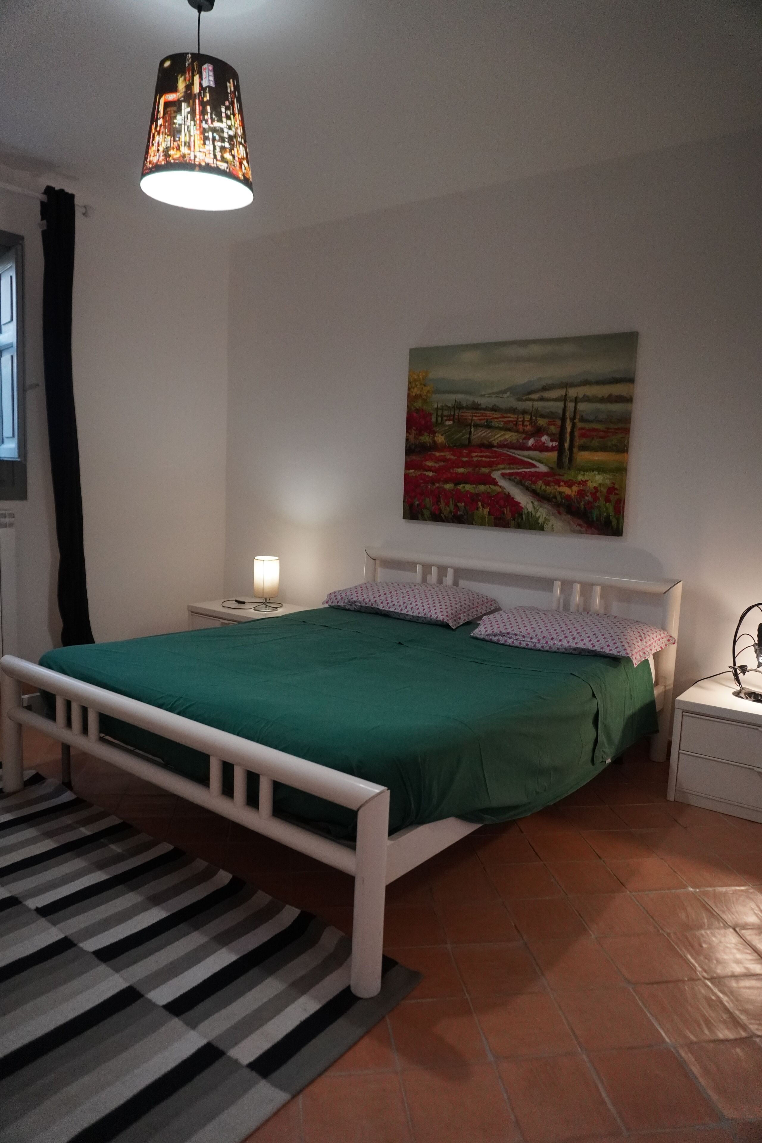 A bedroom with a bed, a nightstand, a lamp, a painting, and a hanging light fixture.